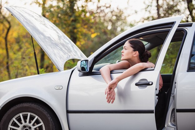 Young woman waits for assistance near her car, which broken down on the road side