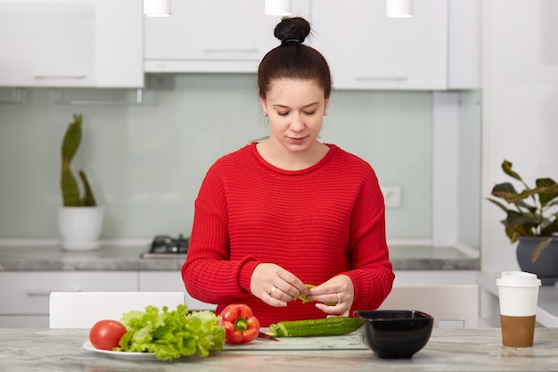 Young woman waits for baby, makes fresh salad at kitchen