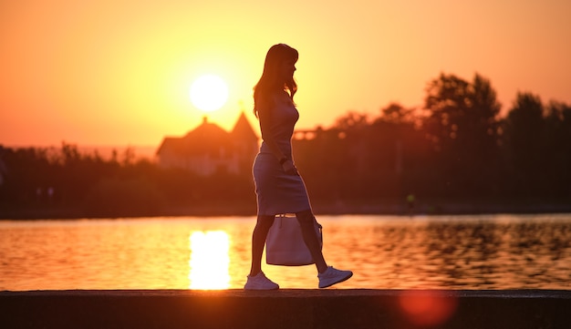 Young woman walking alone on lake shore walkway on warm evening. Solitude and relaxing in nature concept.