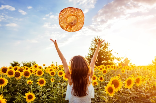 Giovane donna che cammina nel cappello di lancio di fioritura del giacimento del girasole su e che si diverte. vacanze estive