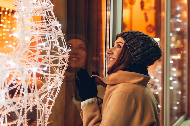 Young woman walking in city and looking at decorated Christmas showcases at night. New year concept