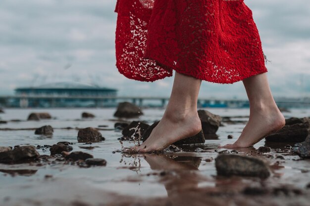 A young woman walks barefoot by the lake in a red dress legs close up