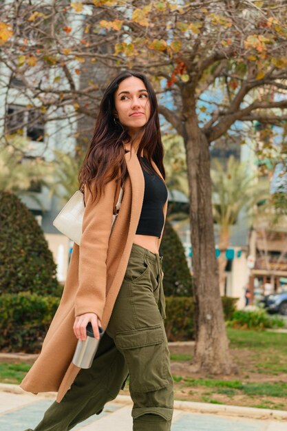 Una giovane donna cammina per strada con abiti casual e un thermos