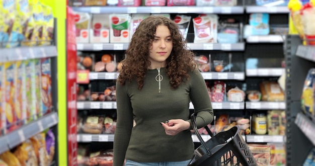 Young woman walks through the supermarket hall with a cart Girl choos the goods in the store and puts them in the basket