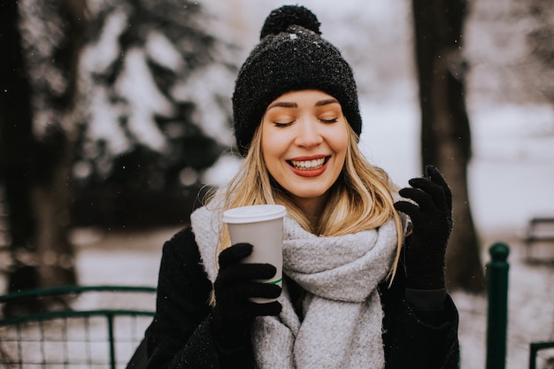 テイクアウトのコーヒー カップと雪の中で楽しんでいる暖かい服装の若い女性