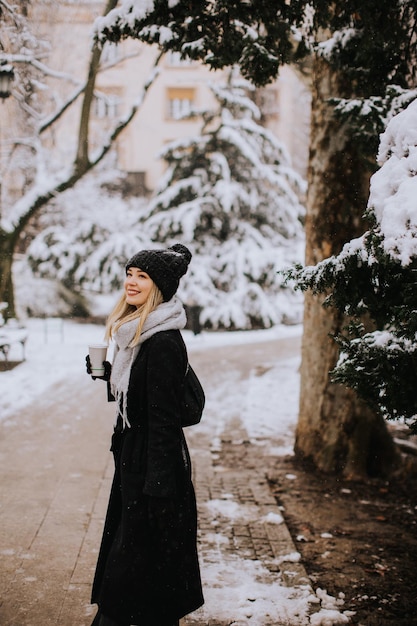 テイクアウトのコーヒー カップと雪の中で楽しんでいる暖かい服装の若い女性