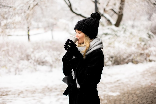 テイクアウトのコーヒー カップと雪の中で楽しんでいる暖かい服装の若い女性