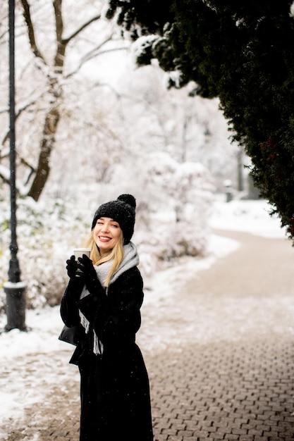 テイクアウトのコーヒー カップと雪の中で楽しんでいる暖かい服装の若い女性