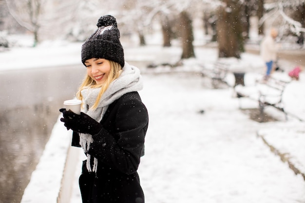 テイクアウトのコーヒー カップと雪の中で楽しんでいる暖かい服装の若い女性