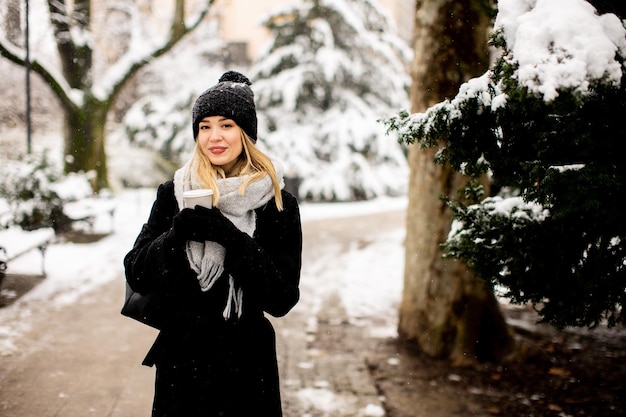 テイクアウトのコーヒー カップと雪の中で楽しんでいる暖かい服装の若い女性