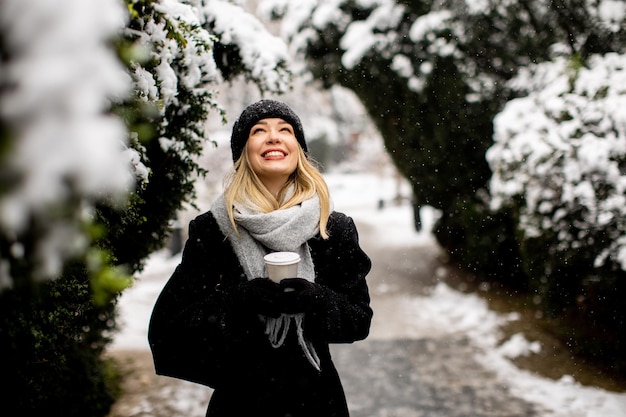 Giovane donna in vestiti caldi che gode nella neve con la tazza di caffè da asporto