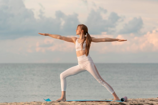 Young woman in the warrior pose outdoors Girl practices yoga at dawn on sea background Yoga at the beach