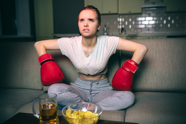 Young woman watch boxing on tv at night. Serious upset model sit on sofa and lean forward. Hands in sport gloves. Beer and chips on table. Alone in dark room. Streamming services.