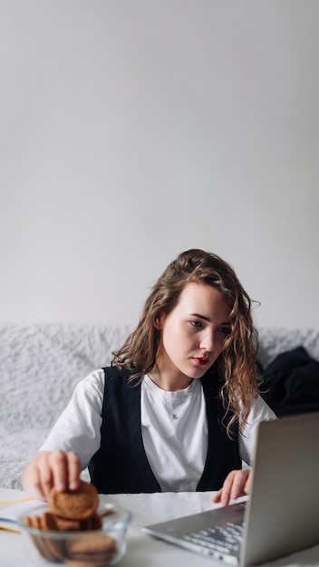 Young woman watching series or podcasts looking at the screen with interest holding cookies in hand sitting at the table with a glass jar and laptop smiling happily Unconscious Eating Disorder