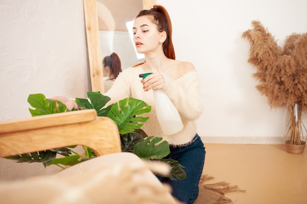 Young woman watering a flower beige interior green monstera 