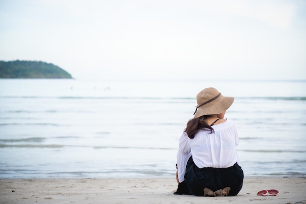 写真 若い女性は海に面したビーチに座って帽子をかぶって、横に眼鏡をかけています。