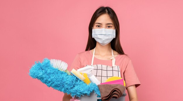 Young woman wear masks to protect disease and apron with holding bucket of cleaning supplies on pink background.