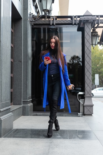 青いコートを着た若い女性がコーヒーを飲みに行き、通りで電話で話している。リアルタイムのミディアムショットをロックダウン