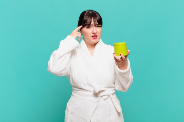 Young woman wearing bathrobe feeling confused and puzzled, showing you are insane, crazy or out of your mind