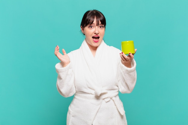 Young woman wearing bathrobe feeling happy, excited, surprised or shocked, smiling and astonished at something unbelievable