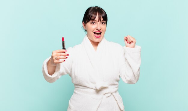 Young woman wearing bathrobe feeling shocked, excited and happy, laughing and celebrating success, saying wow!