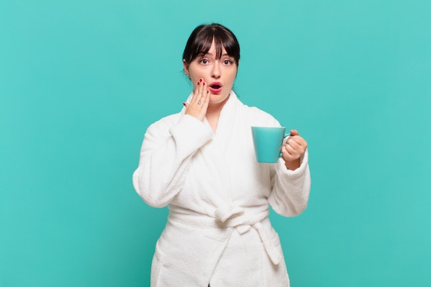 Young woman wearing bathrobe feeling shocked and scared, looking terrified with open mouth and hands on cheeks