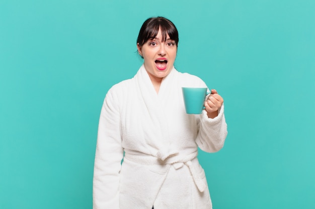 Young woman wearing bathrobe looking happy and pleasantly surprised, excited with a fascinated and shocked expression