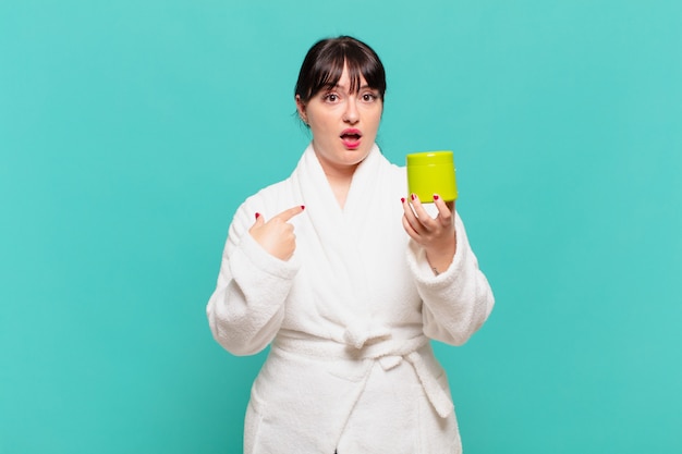 Young woman wearing bathrobe looking shocked and surprised with mouth wide open, pointing to self