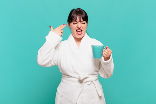 Young woman wearing bathrobe looking unhappy and stressed, suicide gesture making gun sign with hand, pointing to head