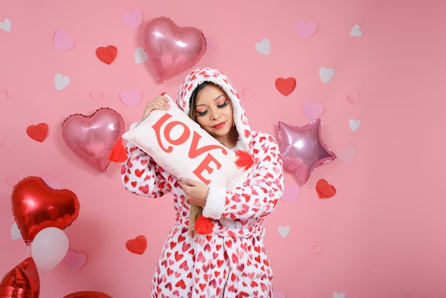 Молодая женщина в халате с сердечками обнимает подушку со словом "любовь"