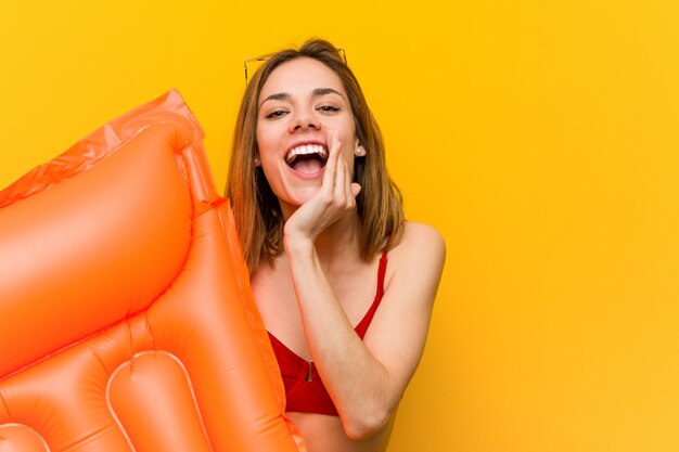 Young woman wearing bikini, holding an air mattress bed shouting excited to front.