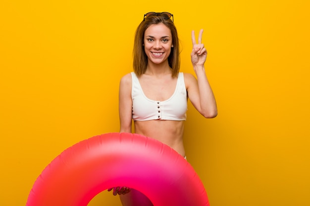 Young woman wearing bikini holding an inflatable donut showing number two with fingers.