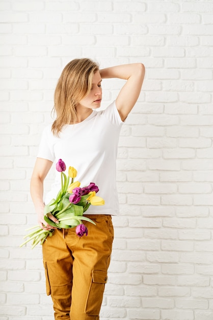 Giovane donna che indossa la maglietta bianca in bianco che tiene i fiori del tulipano che posano contro il muro di mattoni bianchi