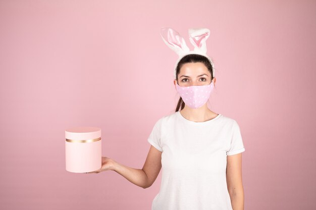 Young woman wearing bunny ears and holding pink gift box. Brunette in protective face mask and white t-shirt on pink background. Easter banner concept. High quality photo