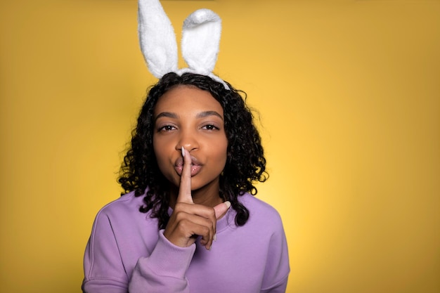 Young woman wearing bunny ears overyellow background asking to be quiet with finger on lips