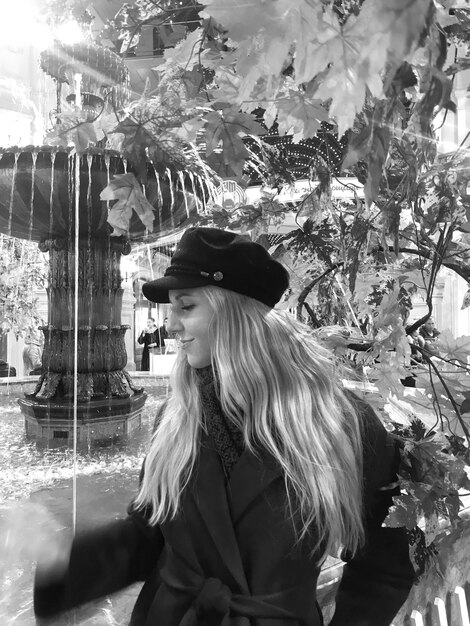 Photo young woman wearing cap and overcoat while standing against fountain in park