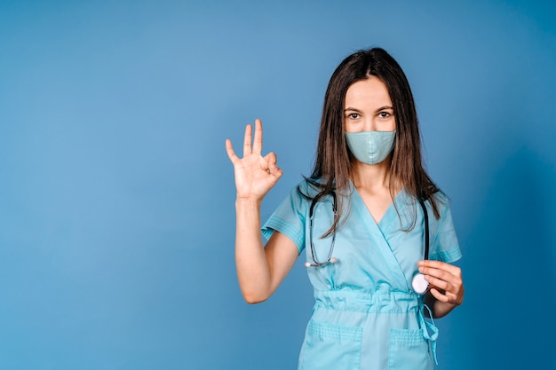 Young woman wearing doctor stethoscope doing ok sign with fingers gesturing excellent symbol wearing the mask