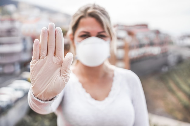 Молодая женщина, носящая лицевую маску и латексные перчатки, показывая жест рукой стоп для предотвращения коронавируса - прекратите распространять концепцию Covid 19 - сфокусируйтесь на руке