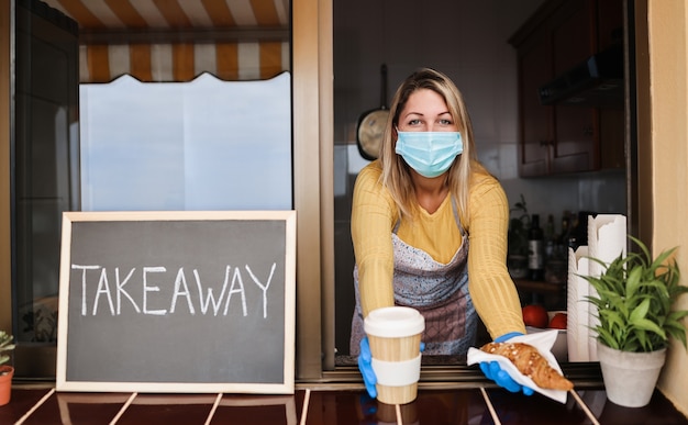 Young woman wearing face mask while serving takeaway breakfast and coffee inside bakery cafe