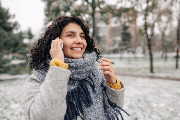 Young woman wearing grey coat and blue scarf talking on the mobile phone at a snowy winter park.