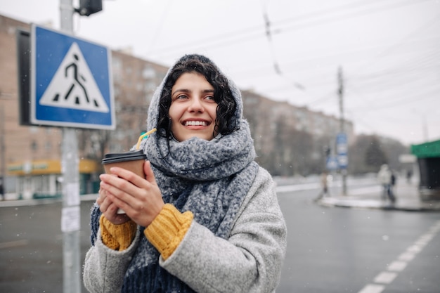 Young woman wearing a grey fashion coat and a blue scarf stands on a crosswalk with a coffee paper cup in her hands.