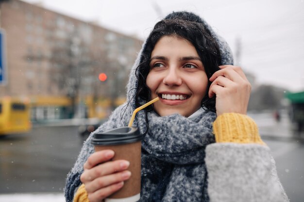 灰色のファッションコートと青いスカーフを身に着けている若い女性は、彼女の手にコーヒーの紙コップを持って横断歩道に立っています。