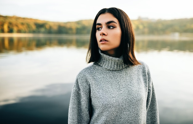 自然の背景にポーズをとって灰色のセーターを着ている若い女性公園の湖に対してポーズをとって片側を見ているかわいい女の子