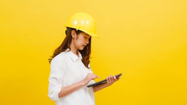 Young woman wearing hardhat using digital tablet against yellow background