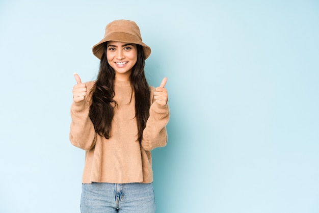 Молодая женщина в шляпе, изолированной на синей стене, улыбается и поднимает палец вверх