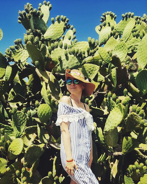 young woman wearing hat and sunglasses while standing against cactus during sunny day