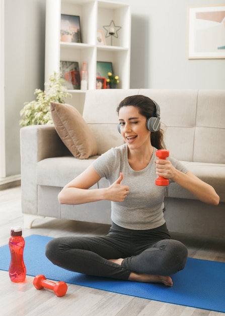 Young woman wearing headphones exercising with dumbbell on yoga mat in front sofa in living room