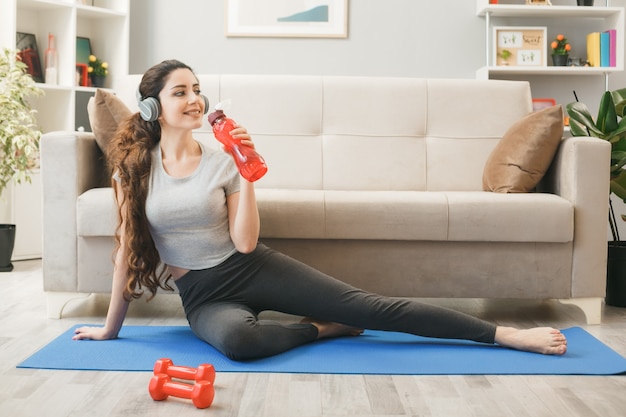 Young woman wearing headphones exercising on yoga mat holding water bottle in front of sofa in living room