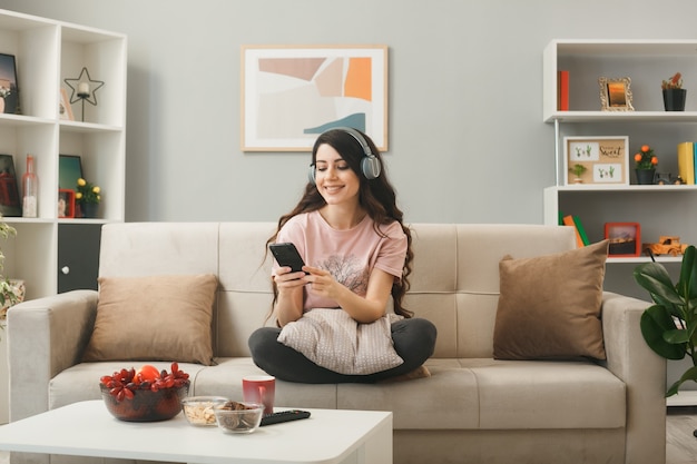 Young woman wearing headphones holding and looking at phone sitting on sofa behind coffee table in living room