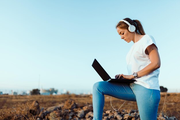 Young woman wearing headphones while using laptop against sky